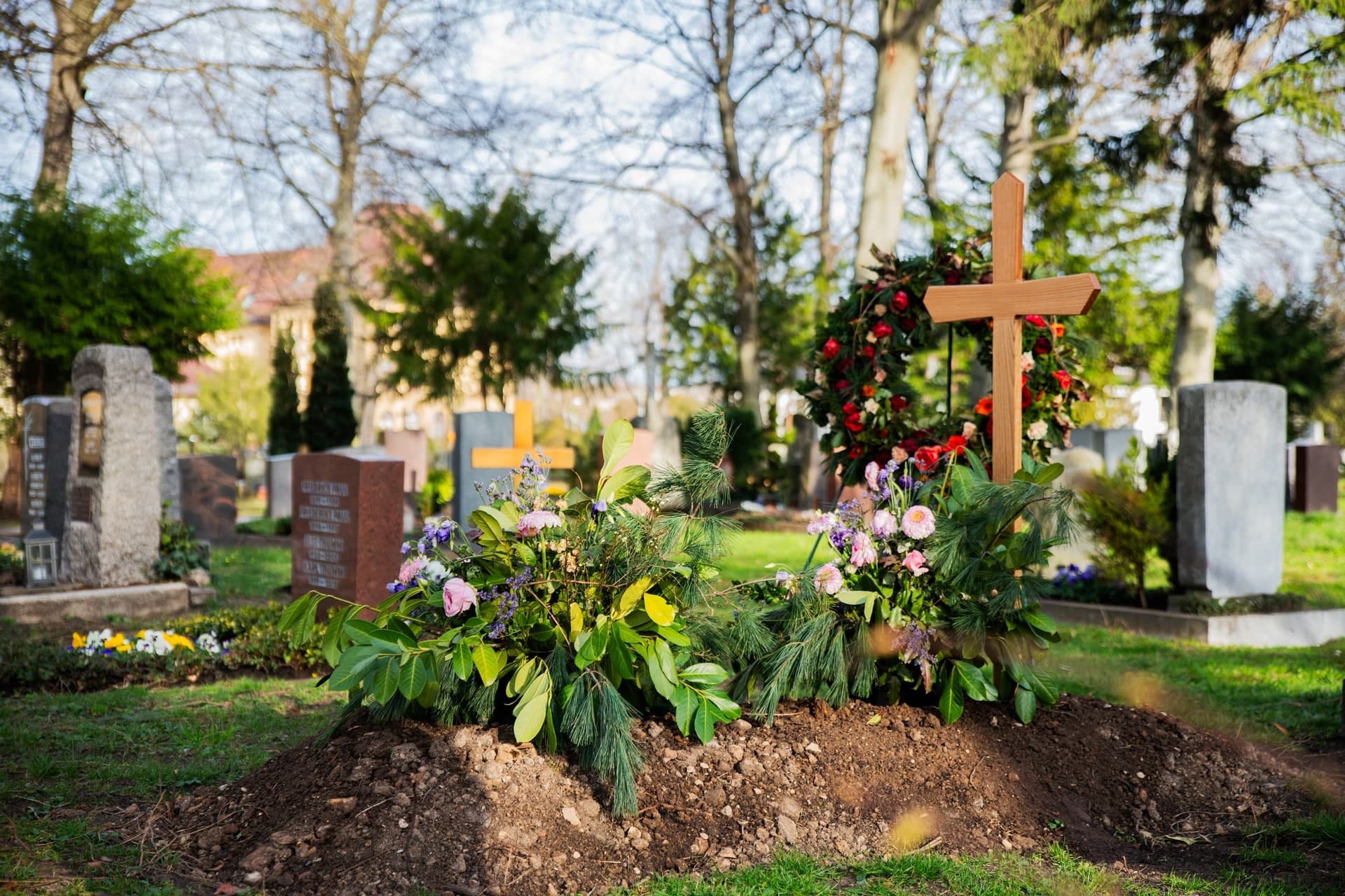 Frisch geschmücktes Grab mit Blumenarrangement und Holzkreuz auf einem sonnigen Friedhof.