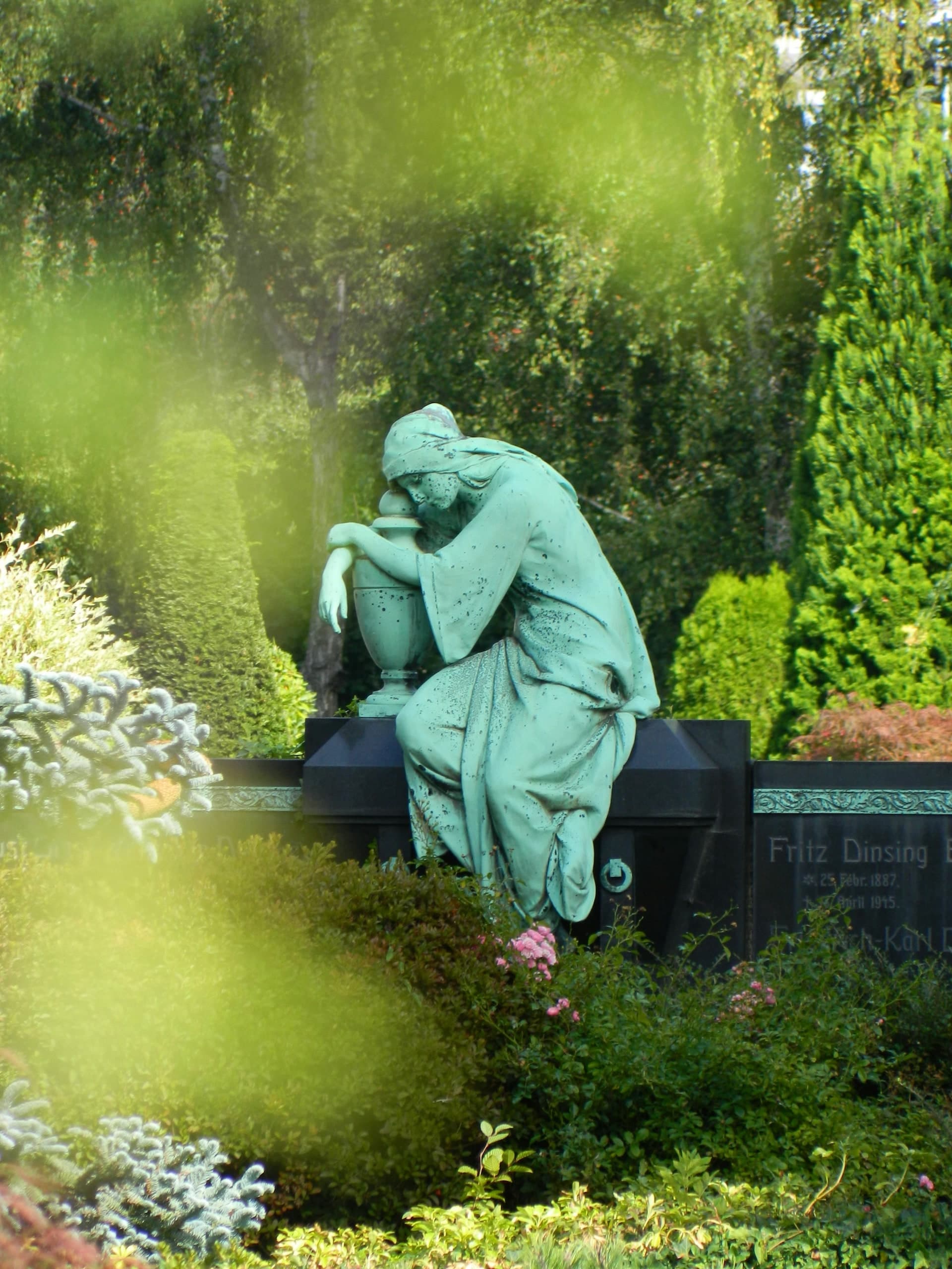 Bronzestatue einer trauernden Frau auf einem historischen Grab inmitten grüner Bepflanzung.