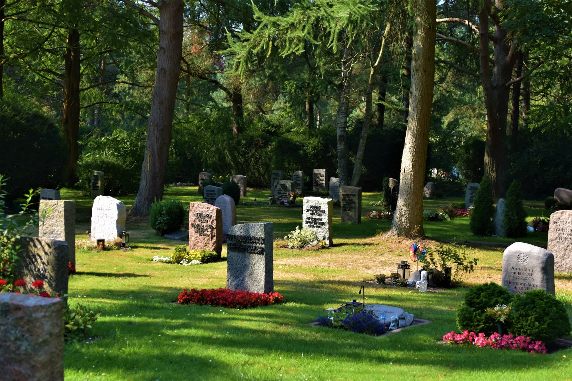 Ruhiger Waldfriedhof mit alten Grabsteinen und blühender Bepflanzung an einem sonnigen Tag.