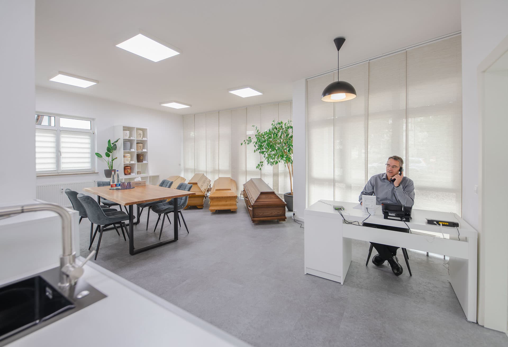 Büro Schwieberdingen – Michael Großhaupt Bestatter Michael Großhaupt telefoniert am Schreibtisch im hellen Büro Schwieberdingen mit Beratungstisch und Särgen