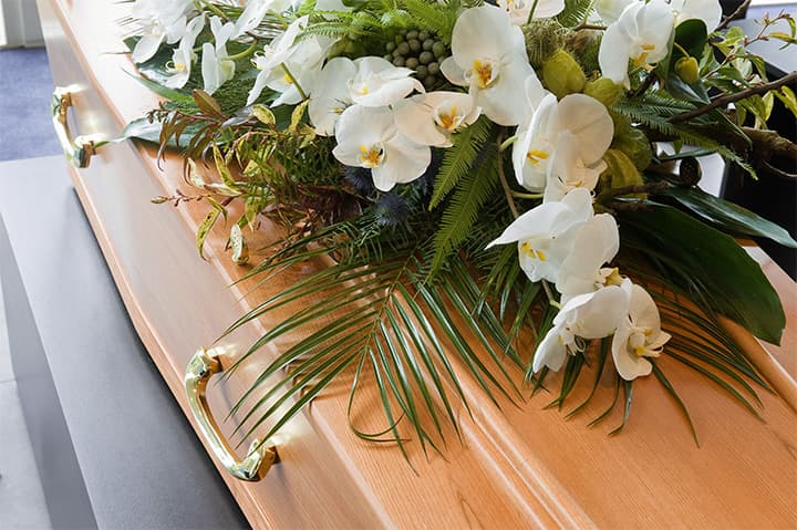 Sarg mit weißem Blumenarrangement Heller Holzsarg mit goldenen Griffen und weißem Blumengesteck aus Orchideen und Grün