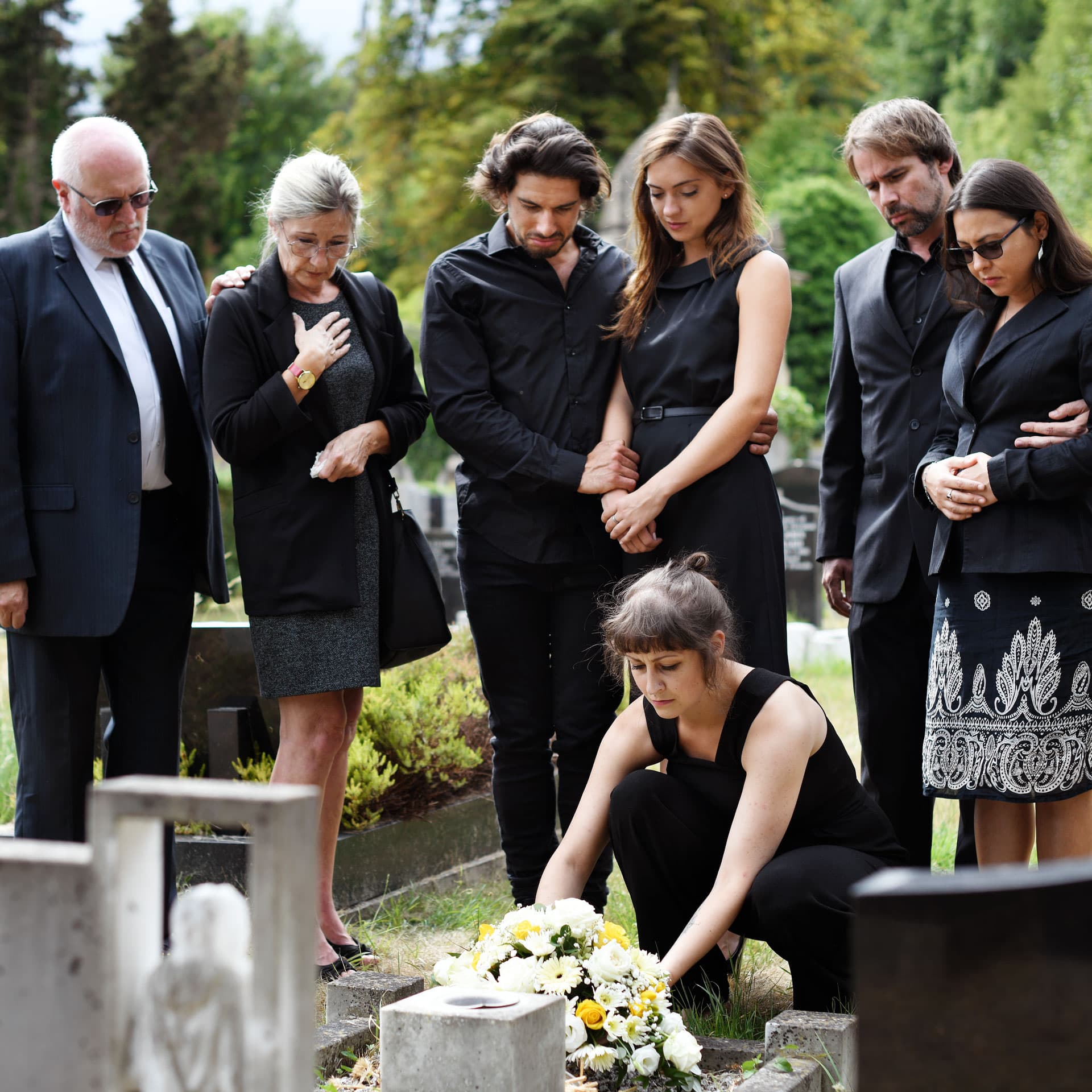 Eine Trauergemeinde in schwarzer Kleidung steht andächtig an einem Grab auf dem Friedhof, während eine Frau einen Blumenstrauß niederlegt. Symbolbild zum Thema angemessenes Verhalten und Fettnäpfchen beim Kondolieren – Ratgeber von Kuhn Bestattungen.